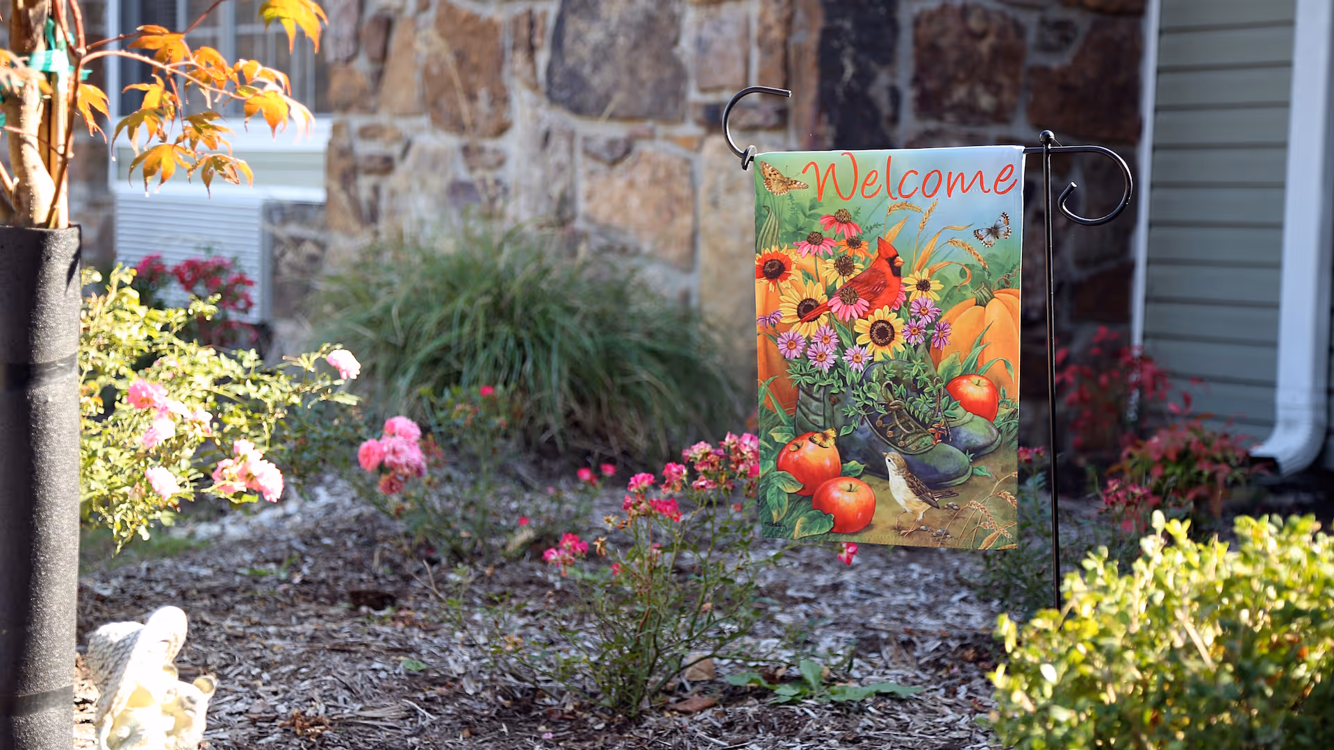 A colorful 'Welcome' garden flag on a metal stand surrounded by flowers and shrubs in front of a stone and siding exterior.