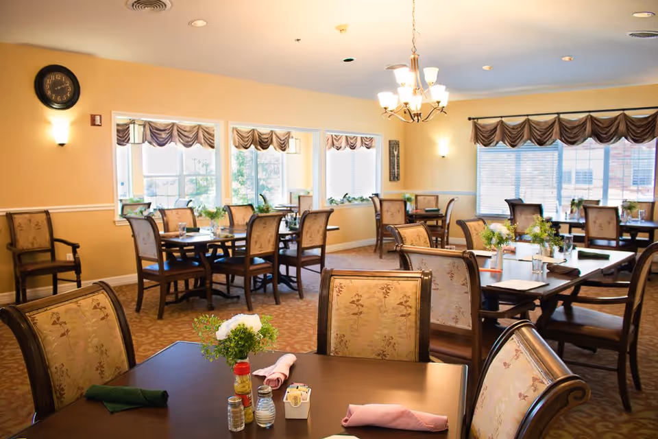 A well-lit dining room with multiple wooden tables and upholstered chairs arranged neatly. Each table has a small floral centerpiece, napkins, and condiments. Large windows with valances allow natural light to fill the room, and a chandelier hangs from the ceiling. A clock and wall sconces are visible on the walls.