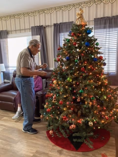 An elderly man decorating a Christmas tree with colorful ornaments and lights in a living room. The room has a brown leather couch, a window with gray curtains, and patterned wallpaper with a decorative border near the ceiling.