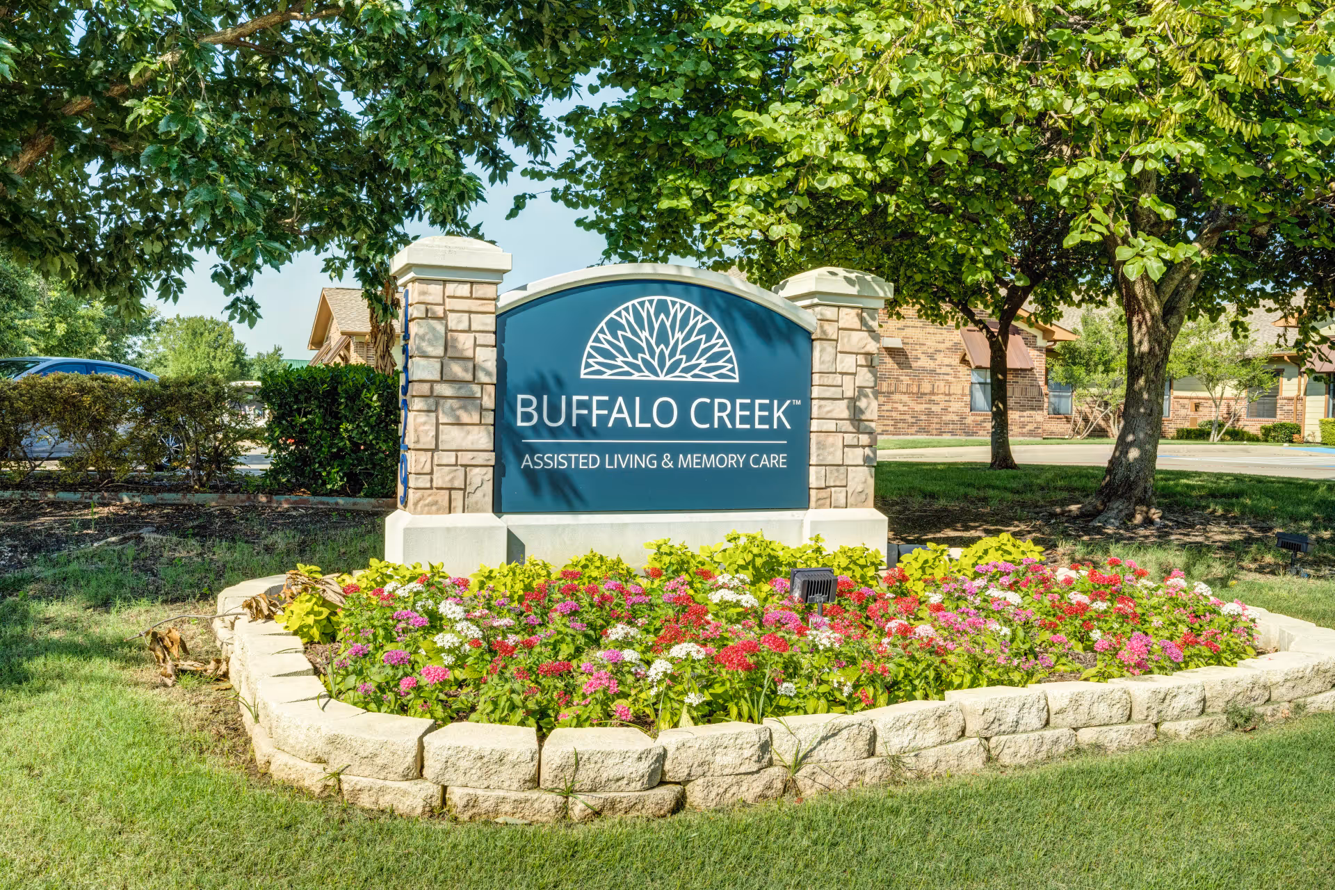 Outdoor view of the Buffalo Creek Assisted Living and Memory Care sign surrounded by a flower bed with colorful flowers and greenery, with trees and part of the building visible in the background.