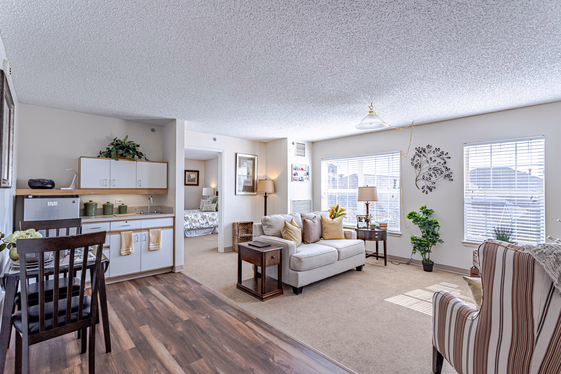 A bright and cozy living area in a senior living facility featuring a small kitchenette with white cabinets and a mini fridge, a dining table with chairs, a beige sofa with multiple pillows, a side table with a lamp, a striped armchair, and large windows with blinds letting in natural light. A bedroom with a bed and framed artwork is visible through an open doorway.