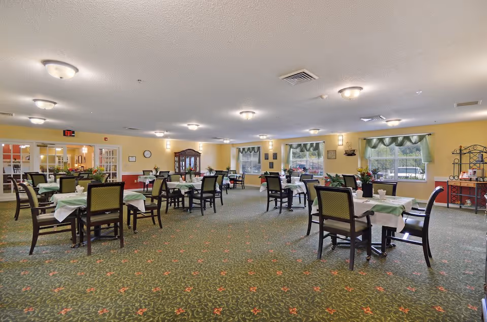 Spacious dining room with multiple tables and chairs set with green tablecloths and floral centerpieces under ceiling lights.