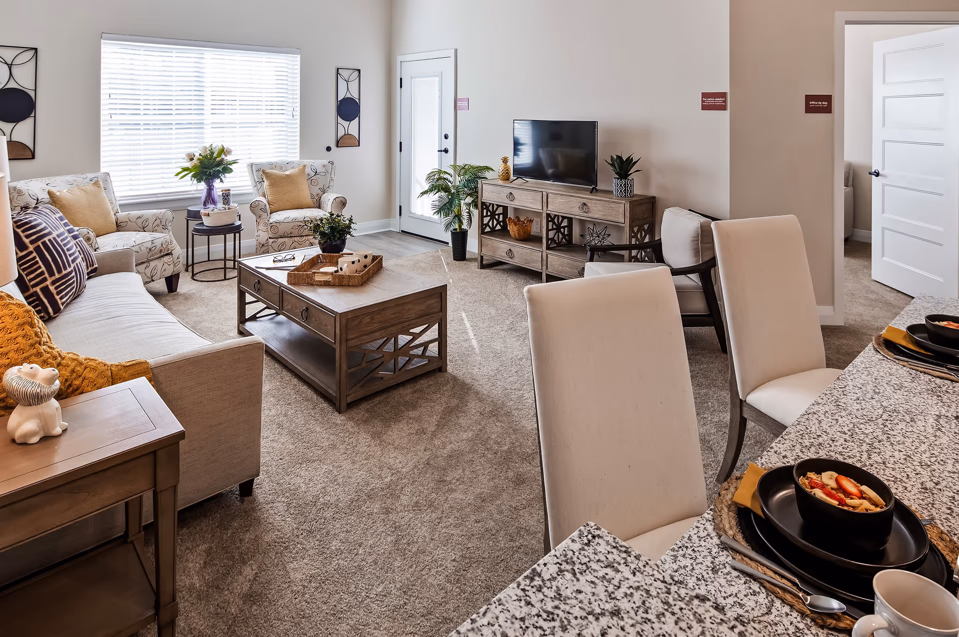 A cozy living room with a beige sofa, two patterned armchairs, a wooden coffee table, and a TV on a wooden stand. There are decorative plants and wall art, a large window with blinds, and a granite countertop with two place settings in the foreground.