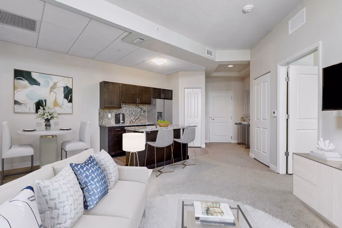 Open-plan living area with a sofa and coffee table facing a kitchen island and dining nook in a modern senior apartment.
