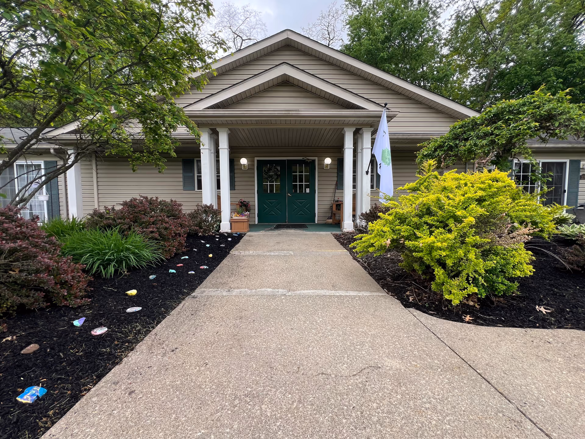 Front entrance of a single-story building with beige siding and green double doors under a covered porch supported by white columns. The walkway leading to the entrance is flanked by landscaped garden beds with various green shrubs and small trees. A white flag is displayed near the entrance.