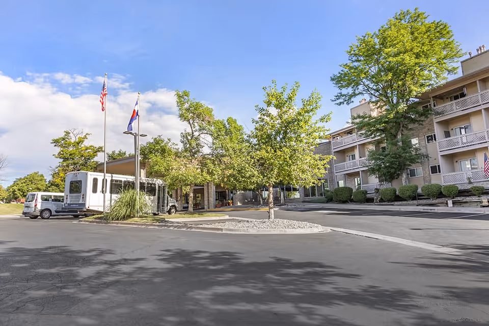 Exterior view of Brookdale Meridian Westland facility showing a parking area with vehicles, two flagpoles with American and state flags, trees, and a multi-story building with balconies under a partly cloudy sky.