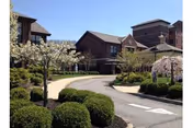 Curved driveway leading to a brick building surrounded by manicured bushes and blooming trees under a clear sky.