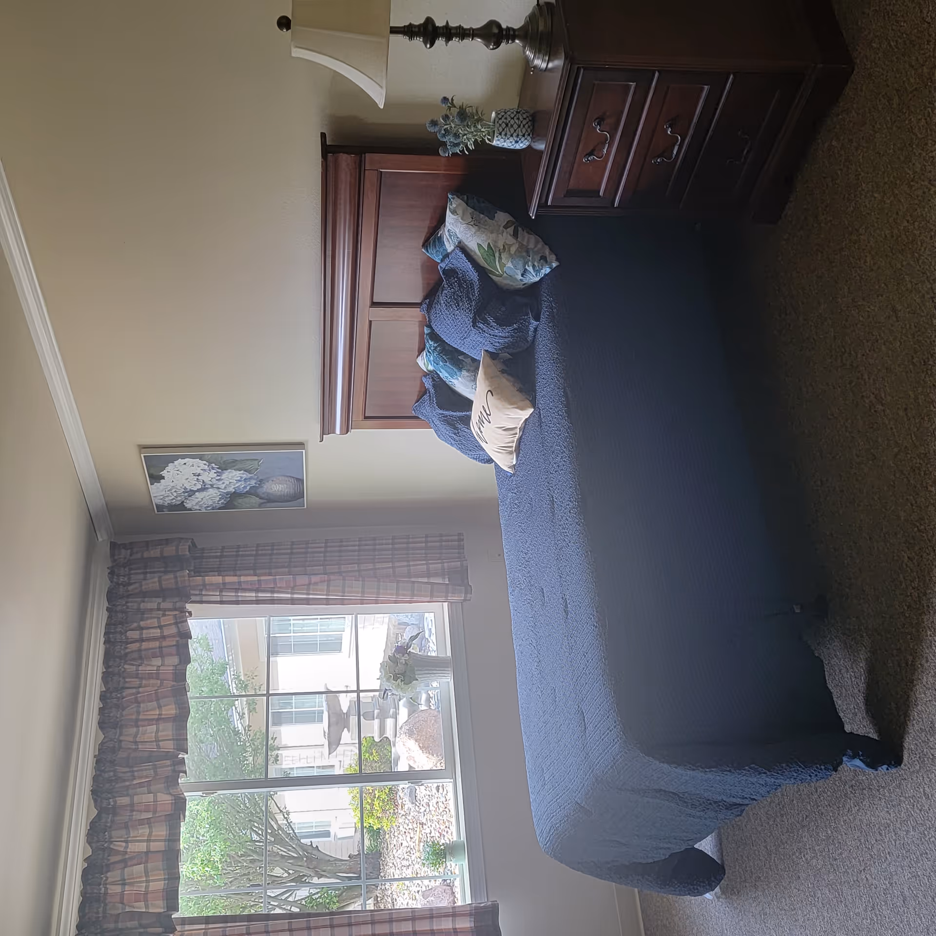 A bedroom with a wooden bed frame and a navy blue quilted bedspread. The bed has several decorative pillows, including one with the word 'home' on it. Next to the bed is a wooden nightstand with three drawers, a table lamp, and a small vase with flowers. A large window with plaid curtains shows an outdoor garden area with rocks, plants, and a fountain. A framed floral painting hangs on the wall above the bed.