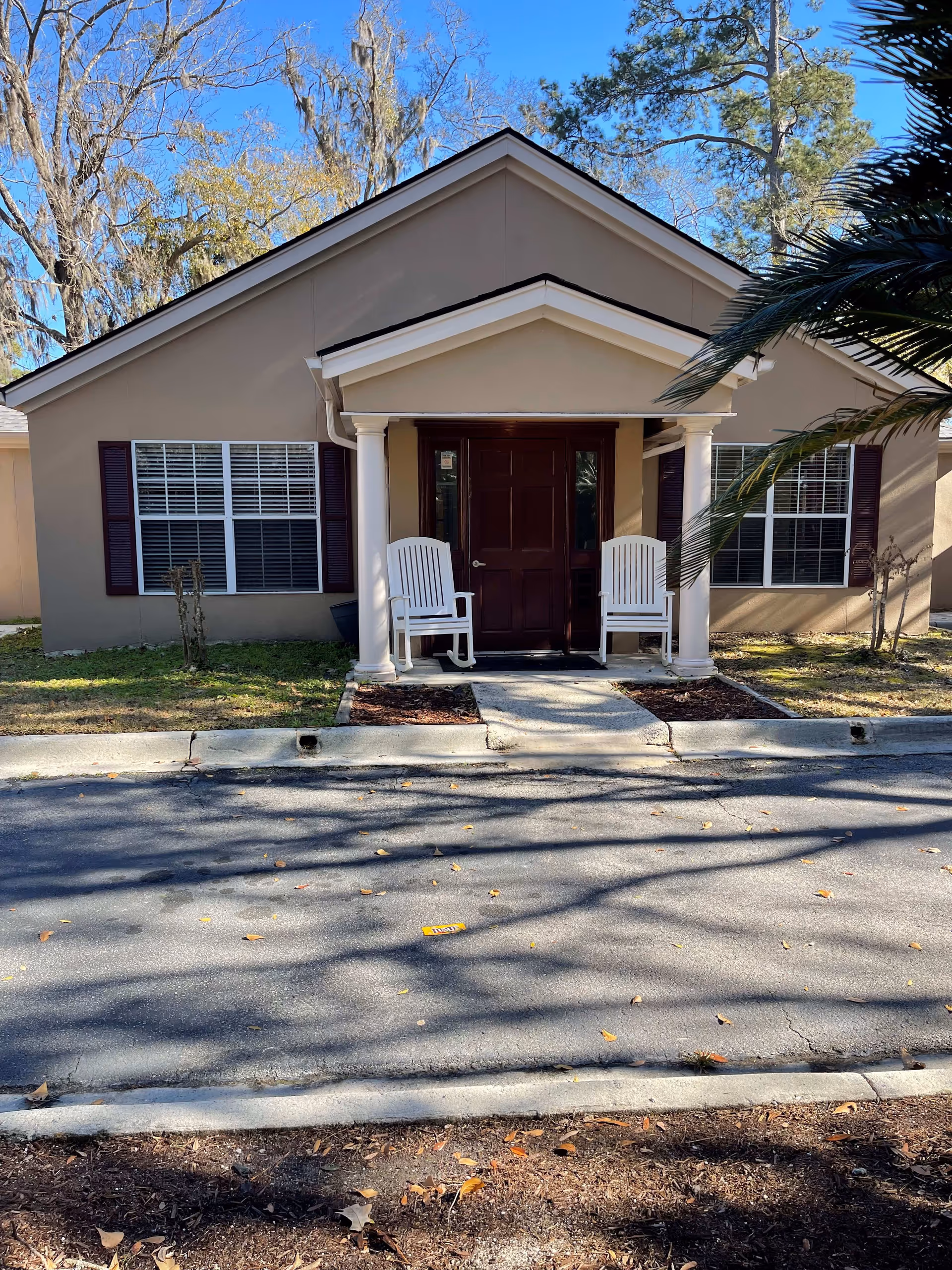 Front exterior view of a single-story beige building with a dark brown door and two white rocking chairs on the small porch. The building has two windows with dark shutters on either side of the door, surrounded by trees and clear blue sky.