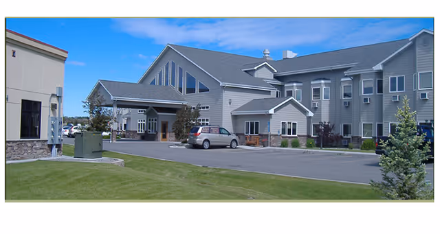 Front exterior of a two-story gray assisted living building with a covered entrance, parked cars and a landscaped lawn under a blue sky.