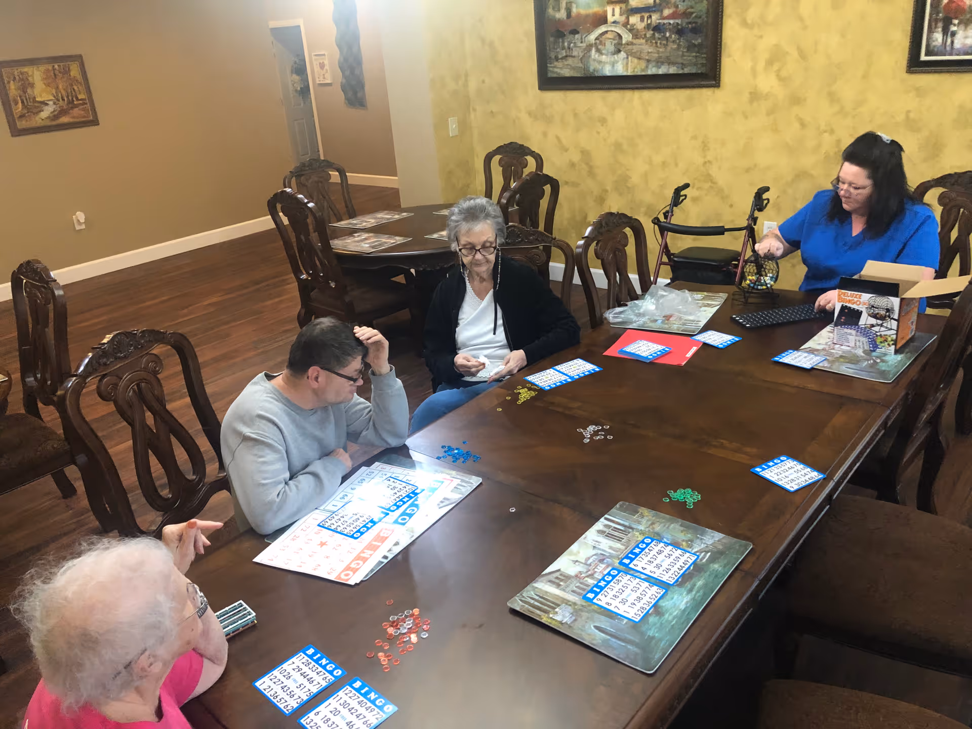 Several residents and a staff member seated around a large wooden table playing bingo in a communal activity/dining room.