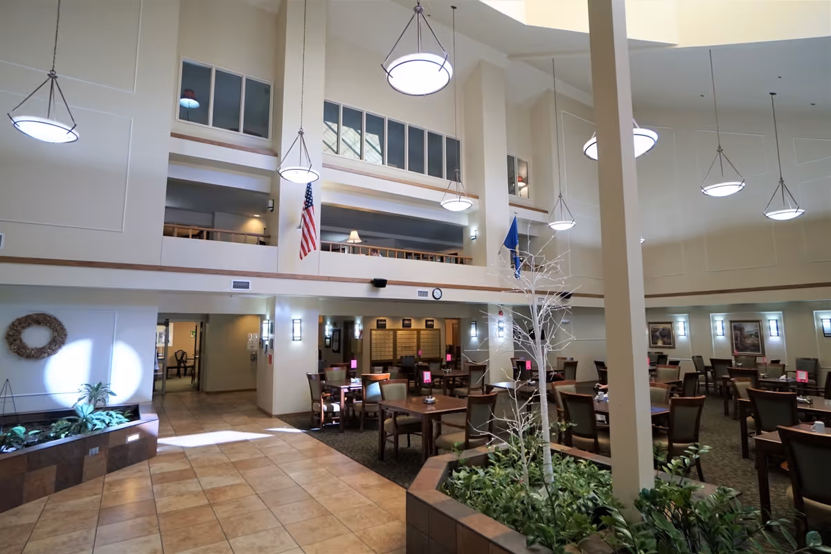 Spacious dining area in a senior living facility with multiple tables and chairs arranged neatly. The room features high ceilings with hanging circular light fixtures, large windows on the upper level, and indoor plants in planters. An American flag and another flag are displayed on the wall, and there is a wreath decoration on the left side. The floor is tiled near the entrance and carpeted in the dining area.