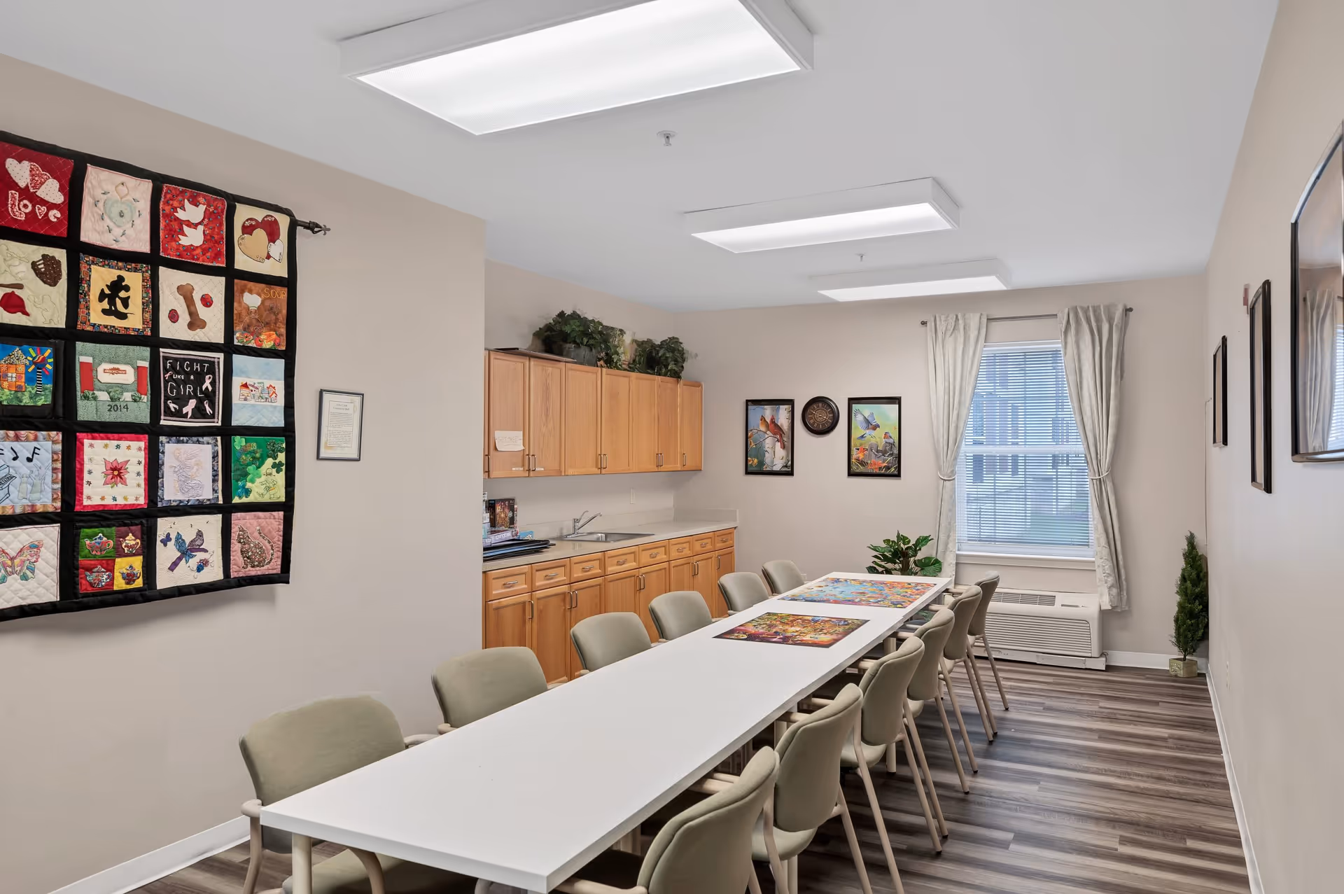 A bright room with a long white table surrounded by beige chairs. On the table are colorful placemats. The room has light-colored walls with framed artwork and a quilt hanging on one wall. There are wooden cabinets with a countertop and a sink along one side of the room. A window with curtains lets in natural light, and the floor is covered with wood-patterned vinyl.