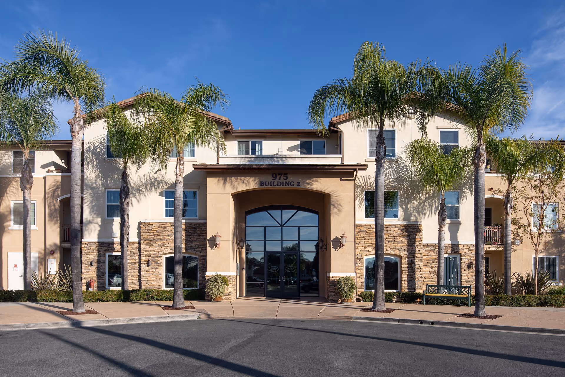 Exterior front view of a three-story building with beige and stone facade, large glass entrance doors under an archway, and several palm trees lining the sidewalk in front.