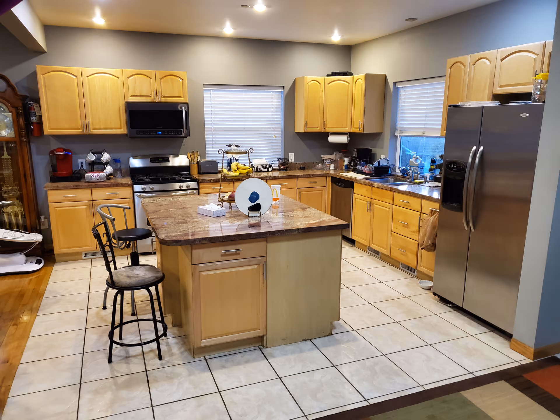 Spacious kitchen with a central island and two bar stools, wooden cabinets, stainless steel appliances, and tiled floor.