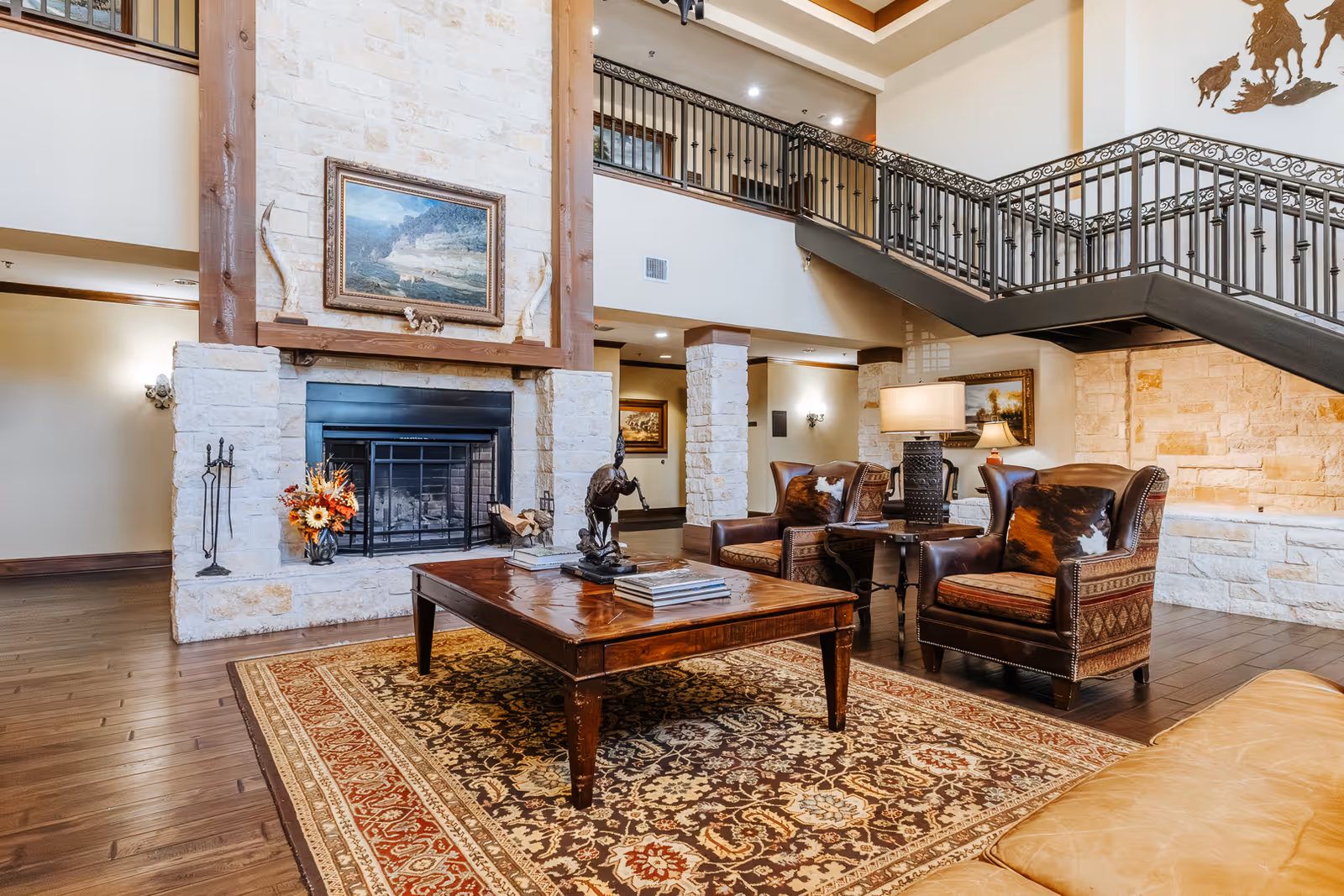 A spacious and elegant living room area with a large stone fireplace as the focal point. The room features a wooden coffee table on a patterned rug, two leather armchairs with decorative pillows, a side table with a lamp, and a staircase with wrought iron railing leading to an upper floor. The walls are light-colored with stone accents, and there is framed artwork above the fireplace and on the walls.