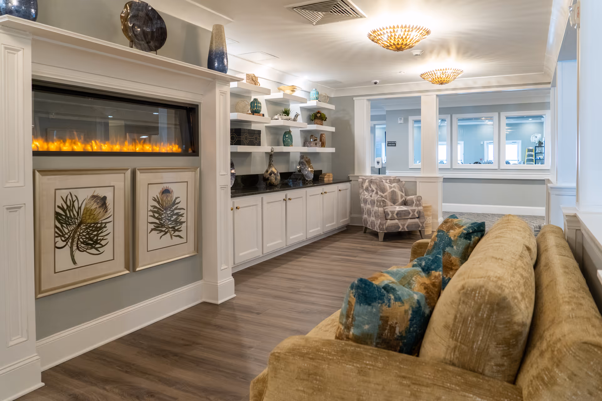 A cozy living room area in Arbor Ridge at Huntersville featuring a beige sofa with colorful cushions, a patterned armchair, a modern electric fireplace with framed botanical artwork below it, white built-in shelves and cabinets decorated with vases and plants, and two ceiling light fixtures.