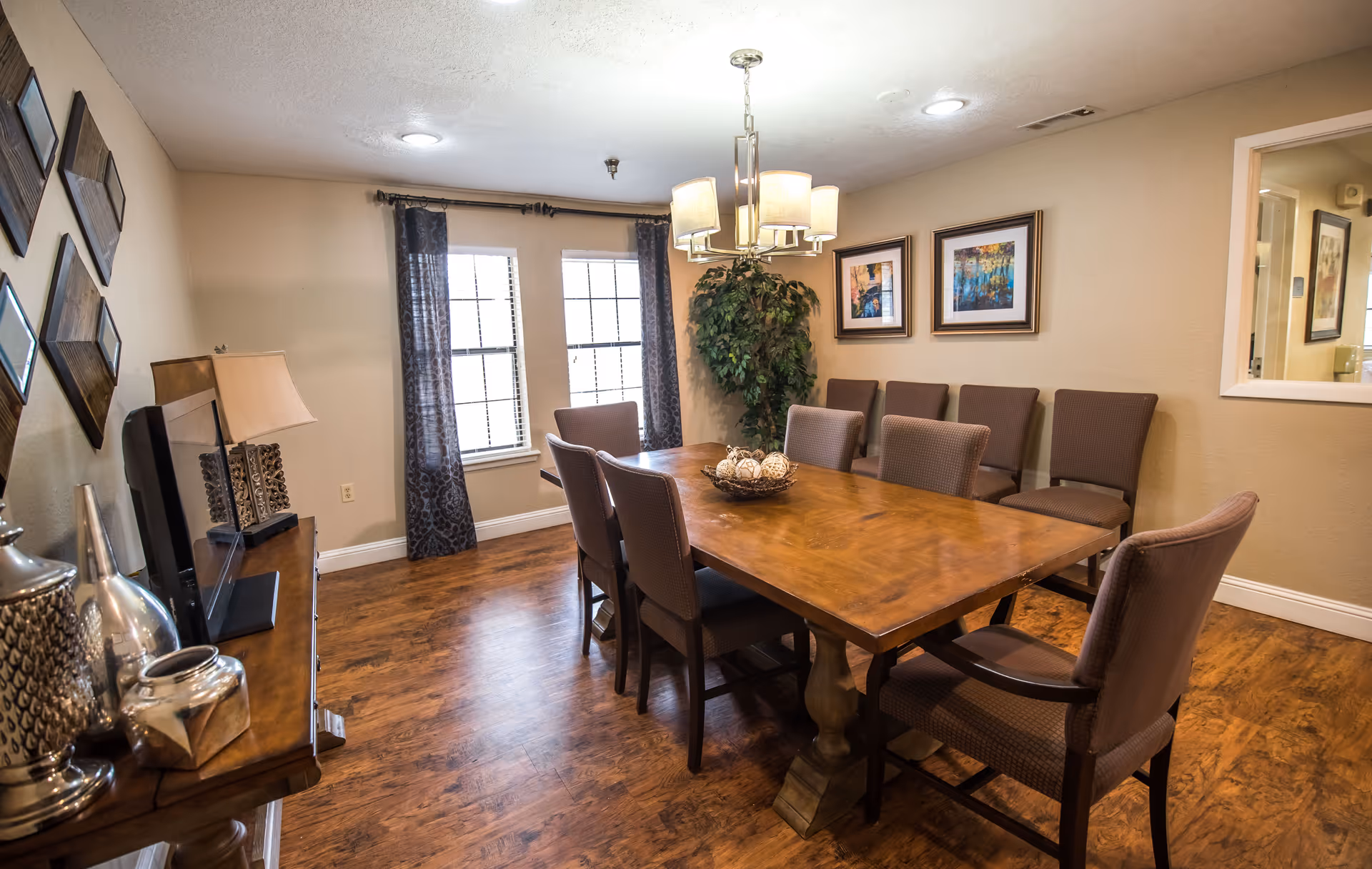 A dining room with a large wooden table surrounded by eight upholstered chairs. The room has wooden flooring, two windows with dark curtains, a chandelier hanging above the table, framed artwork on the walls, and a decorative plant in the corner. A side table with decorative items and a lamp is visible on the left side.