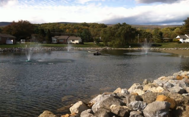 A pond with three water fountains surrounded by rocks in the foreground and residential buildings with trees and hills in the background under a partly cloudy sky.