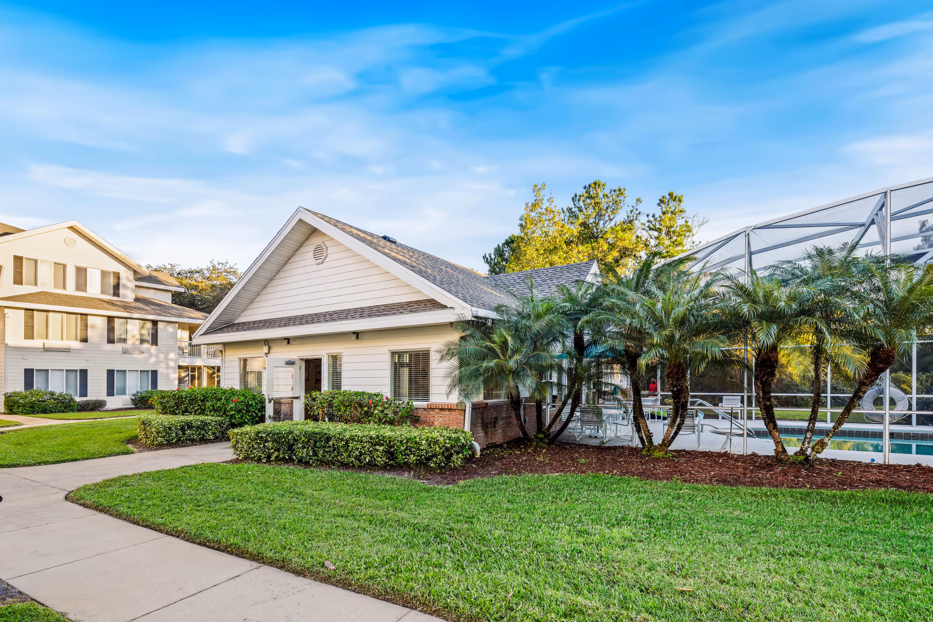 Exterior view of a senior living community building with a well-maintained lawn, palm trees, and a screened-in swimming pool area under a blue sky.