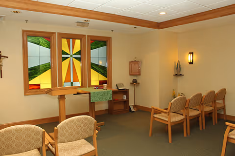 A small chapel or meditation room with wooden chairs arranged in rows facing a wooden podium. Behind the podium is a wall with three stained glass windows featuring a colorful geometric design. The room has beige walls, a green carpet, and soft lighting from wall sconces. There is a small statue on a shelf and a wooden cabinet on the right side.