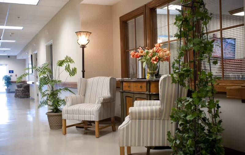 A well-lit hallway in a senior living facility with two striped armchairs, a wooden side table with a vase of flowers, a tall floor lamp, and several potted plants. The hallway has beige walls and a window with blinds looking into another room.
