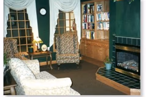 A cozy common living room with upholstered sofa and armchairs, a tiled fireplace, built-in bookshelves, side table with lamp, and draped windows.