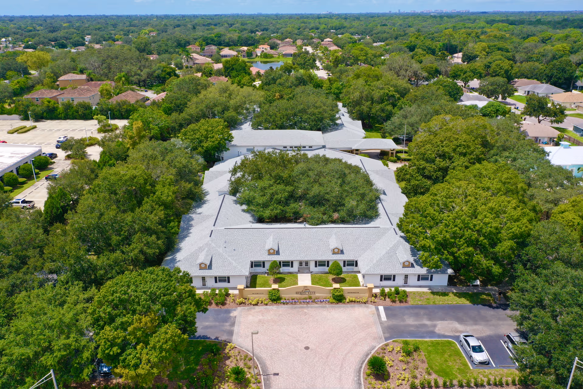 Aerial view of a senior living facility named Heron East surrounded by dense green trees and residential houses. The building has a light gray roof and white walls, with a circular driveway and parking spaces in front. The area is lush with greenery and well-maintained landscaping.