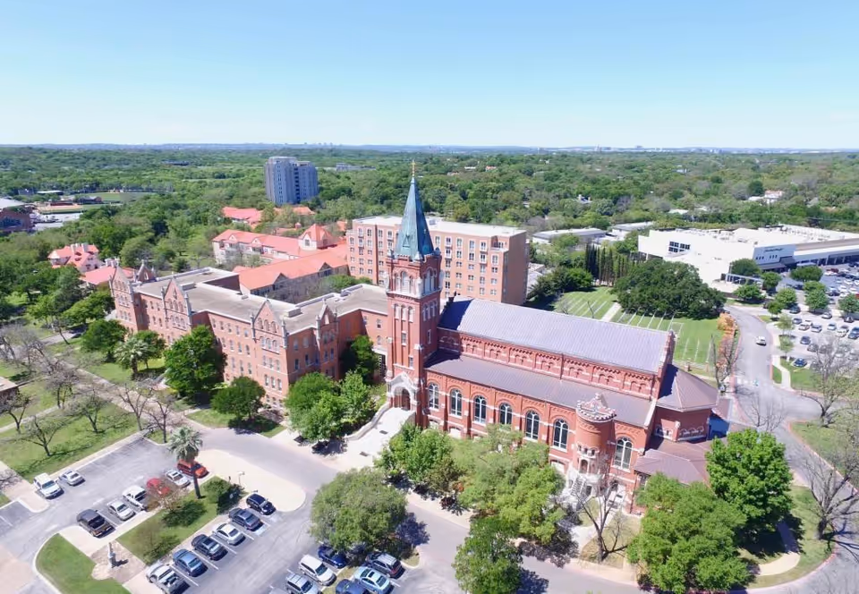 Aerial view of a large historic red-brick campus with a tall central steeple, parking lot, and surrounding trees.