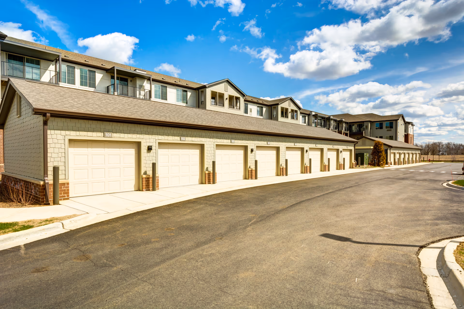 Long row of garage doors facing a paved driveway with a multi-story residential building behind under a blue sky.