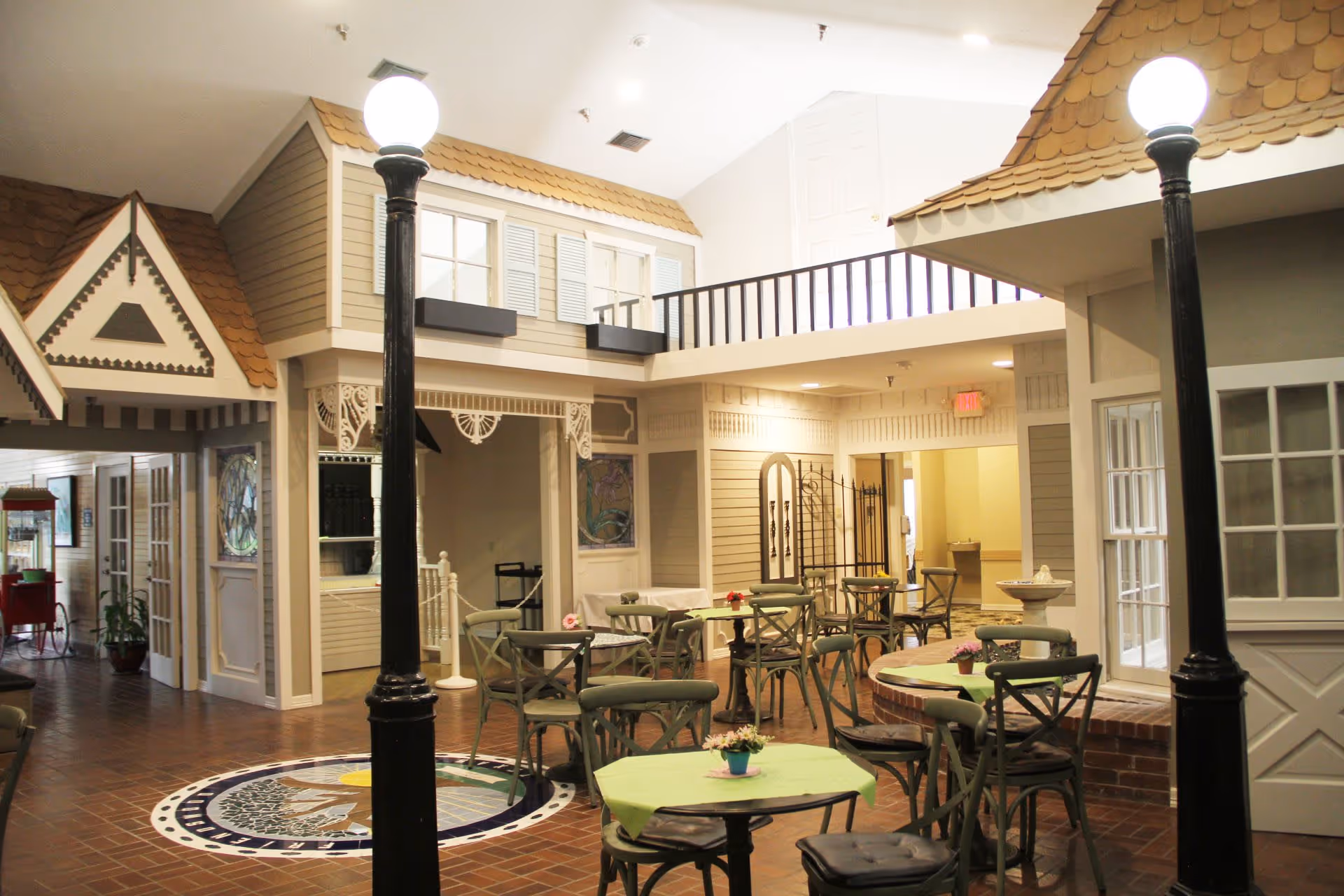 Indoor seating area designed like a small village street with tables and chairs arranged for dining or socializing. The space features house-like facades with windows, shutters, and decorative trim, along with two black street lamps with round white lights. The floor is tiled with a circular mosaic design in the center.