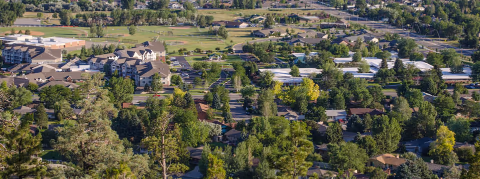 Aerial view of a suburban area with a mix of residential houses, trees, and a large multi-story building complex surrounded by greenery and parking lots.