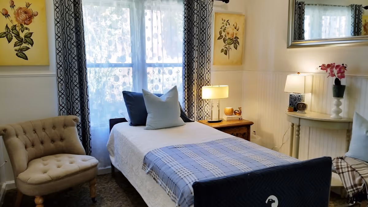 A cozy bedroom with a single bed covered in a white blanket and a blue plaid throw. Two pillows, one dark blue and one light blue, are placed on the bed. To the left of the bed is a beige tufted armchair. Behind the bed is a window with sheer white curtains and patterned drapes. On the right side, there is a wooden nightstand with a lamp and decorative items, and a small white table with another lamp, a clock, and a pink orchid. Floral paintings hang on the walls, and a large mirror is mounted above the white table.