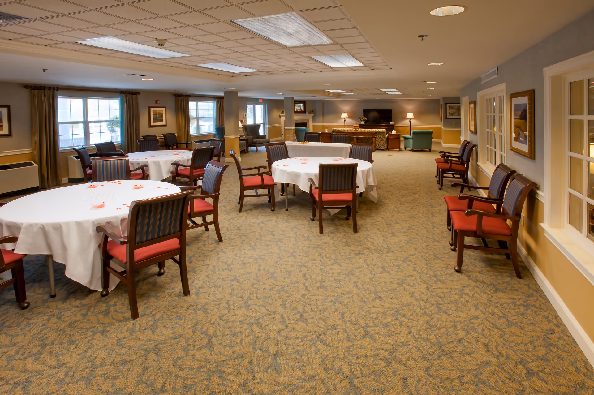 A spacious senior living dining room with round tables covered in white tablecloths and surrounded by wooden chairs with red cushions. The room has large windows with curtains, framed artwork on the walls, and a carpeted floor. In the background, there is a seating area with armchairs, a fireplace, and a television.