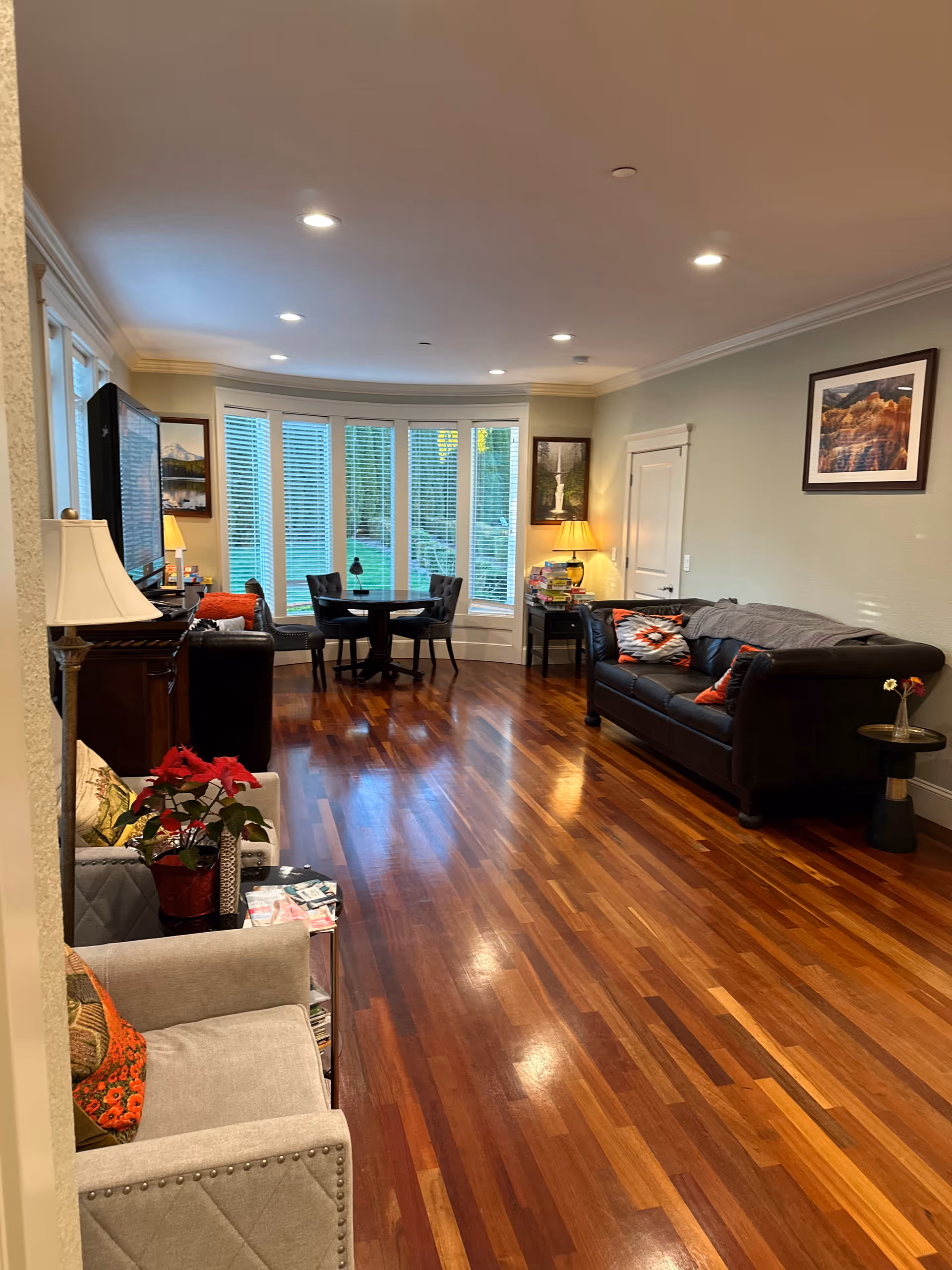 A spacious living room with polished wooden floors, a black leather sofa with colorful cushions, a small round table with four chairs near a large bay window with white blinds, a beige armchair with a red poinsettia plant beside it, framed artwork on the walls, and warm lighting from table lamps.