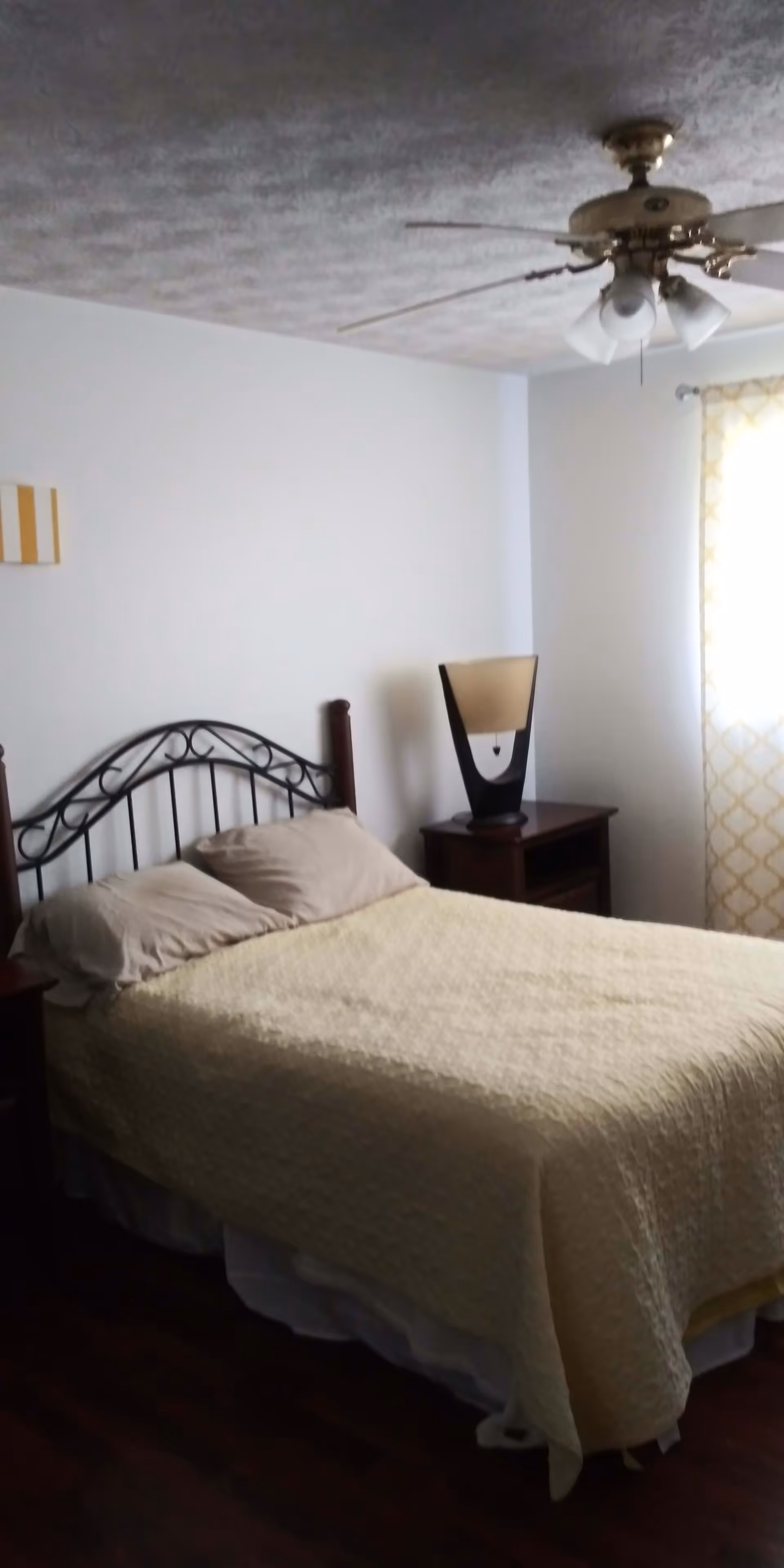 A bedroom with a bed covered in a light yellow quilt and two beige pillows. The bed has a black metal headboard with a decorative design. There is a wooden nightstand next to the bed with a modern lamp on it. A window with yellow and white patterned curtains is on the right side of the room. A ceiling fan with lights is mounted on the ceiling.