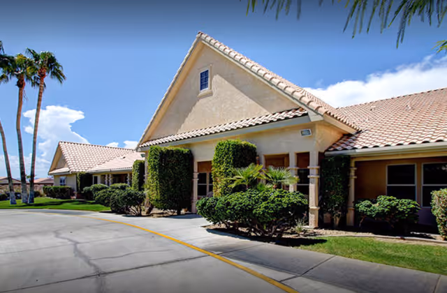 Single-story stucco senior living building with a tiled roof, trimmed bushes and palm trees along a curved driveway under a blue sky.