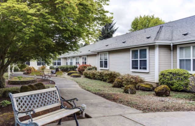A single-story senior living building with a paved walkway, benches, and landscaped shrubs and trees.