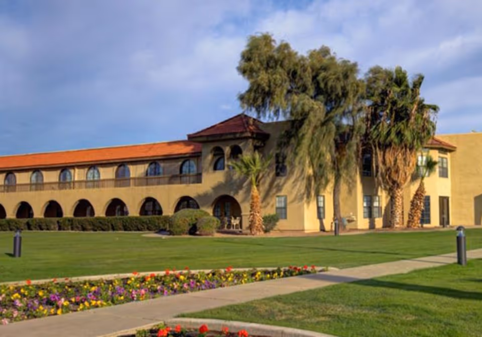 Tan Spanish-style senior living building with arched windows, palm trees, and a manicured lawn