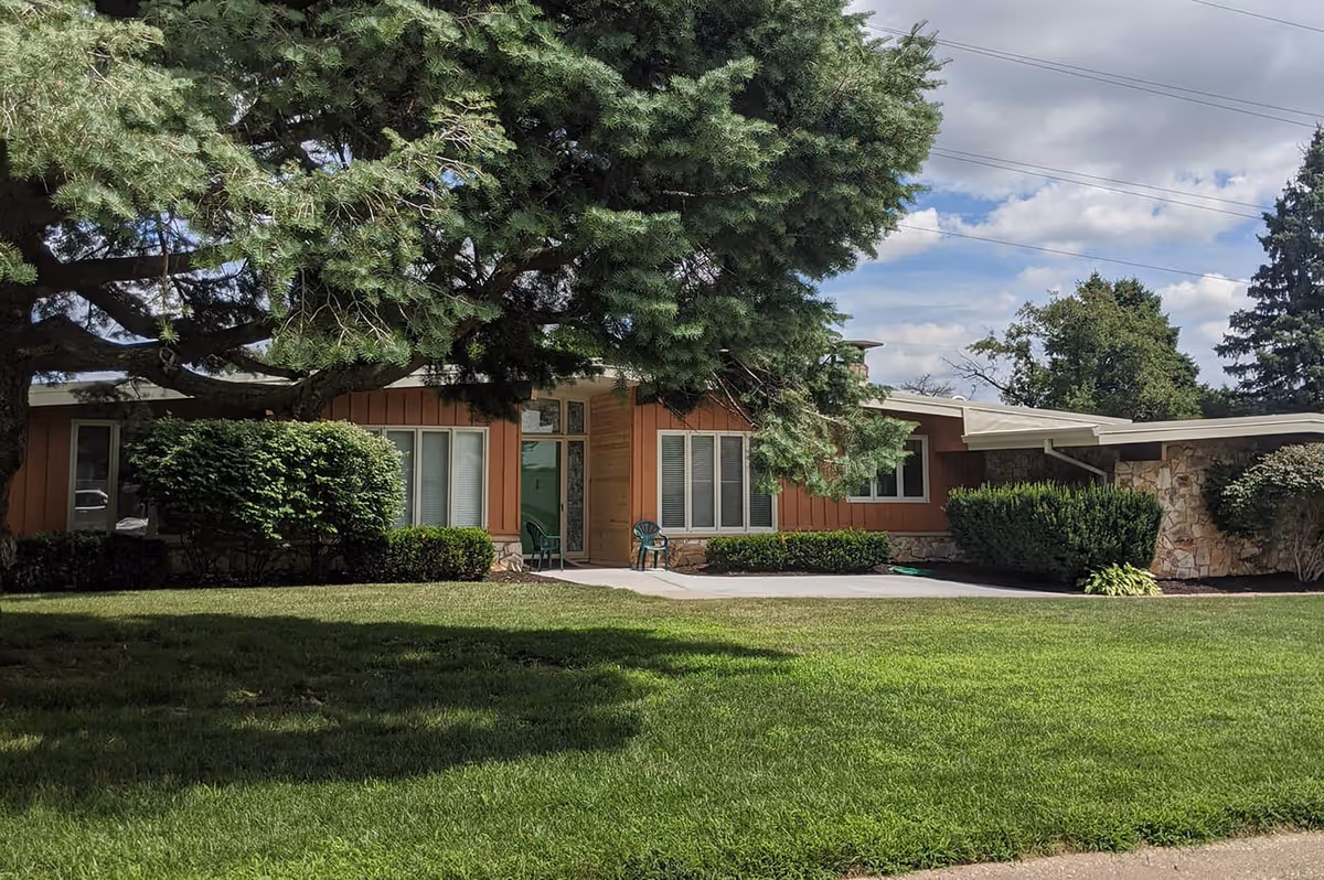 Single-story building with brown siding and stone accents, surrounded by green bushes and a large tree in front, with a well-maintained lawn under a partly cloudy sky.