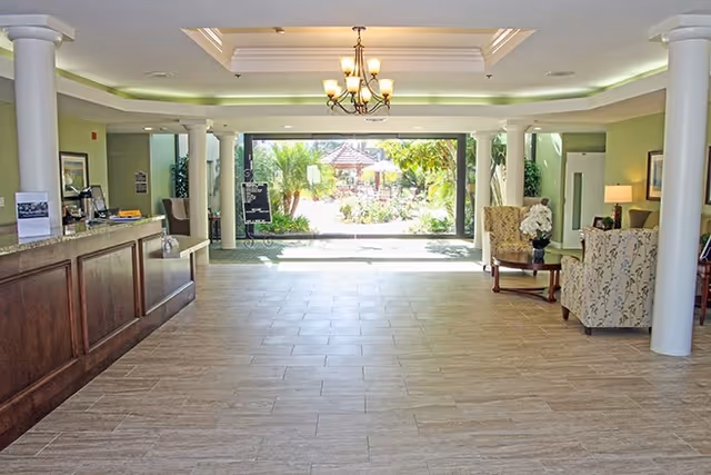 Spacious and well-lit lobby area with a wooden reception desk on the left, comfortable seating with patterned armchairs and a small table on the right, white columns, a chandelier hanging from the ceiling, and large glass doors at the far end opening to a garden with greenery and a gazebo.