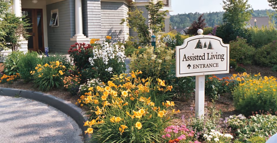 A landscaped garden area with colorful flowers and shrubs in front of a building. A white sign with green tree illustrations reads 'Assisted Living ENTRANCE' with an arrow pointing forward. The building has gray siding and a wooden door partially visible on the left side.