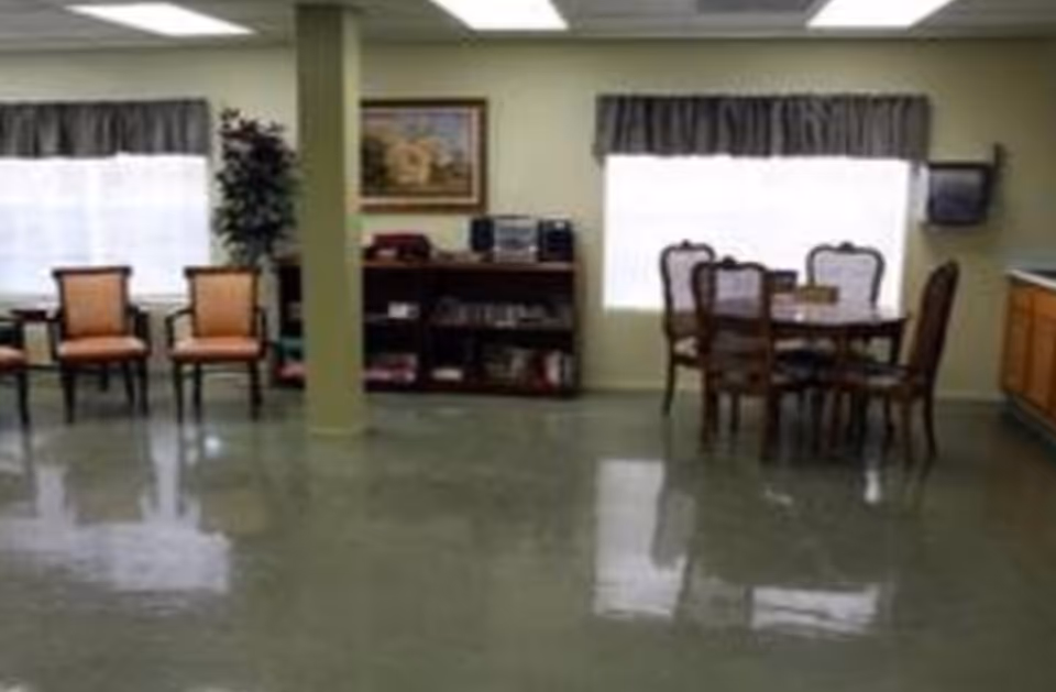 A common area in a senior living facility with several chairs arranged along the left wall, a small bookshelf with a stereo and books in the center, and a dining table with six chairs near a window with curtains. The floor is shiny and reflective.