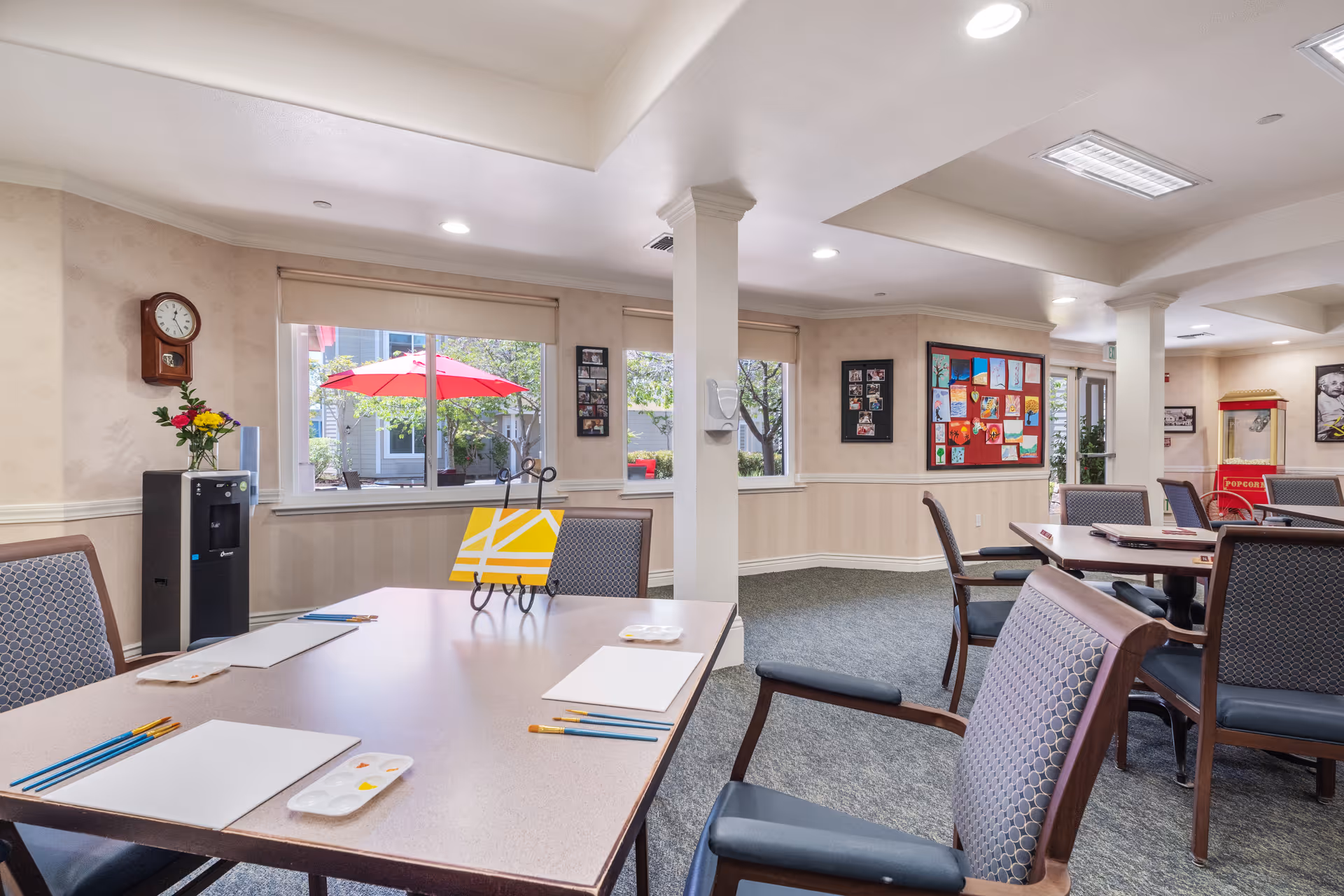 A bright and spacious activity room in a senior living facility with tables and chairs arranged for painting activities. The tables have blank canvases, paint palettes, and paintbrushes set up. There are windows letting in natural light, a water cooler, a wall clock, and colorful artwork displayed on the walls. Outside the windows, a red patio umbrella is visible.