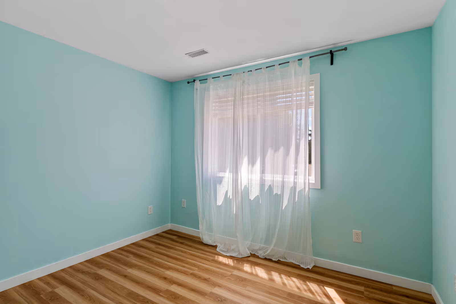 Empty room with light turquoise walls, a window covered with sheer white curtains, and wood-patterned flooring.