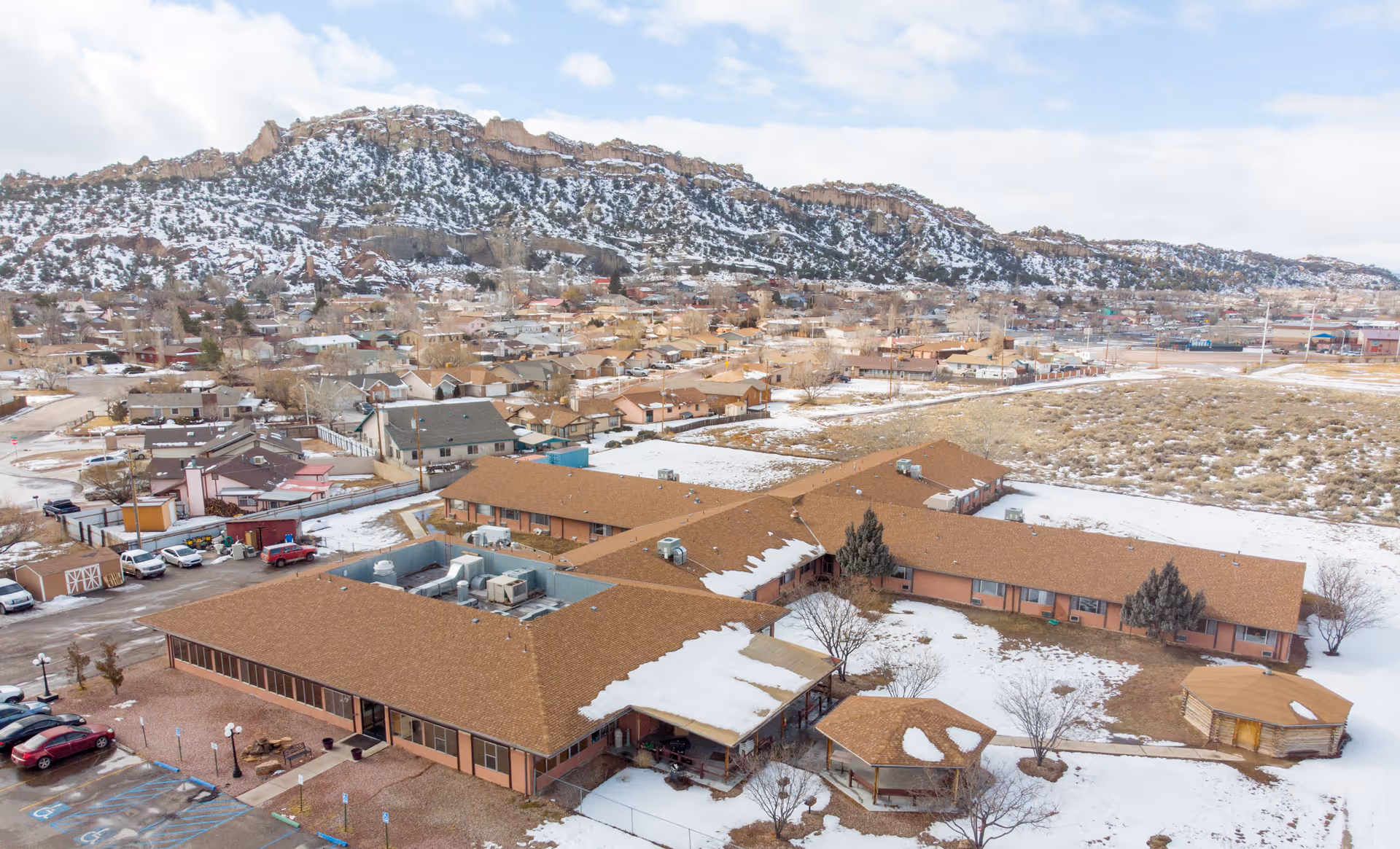 Aerial view of Red Rocks Care Center, a single-story building with a brown roof partially covered in snow. The facility is surrounded by a parking lot with several cars, some trees, and open land with patches of snow. In the background, there is a residential neighborhood and a rocky, snow-dusted mountain range under a partly cloudy sky.