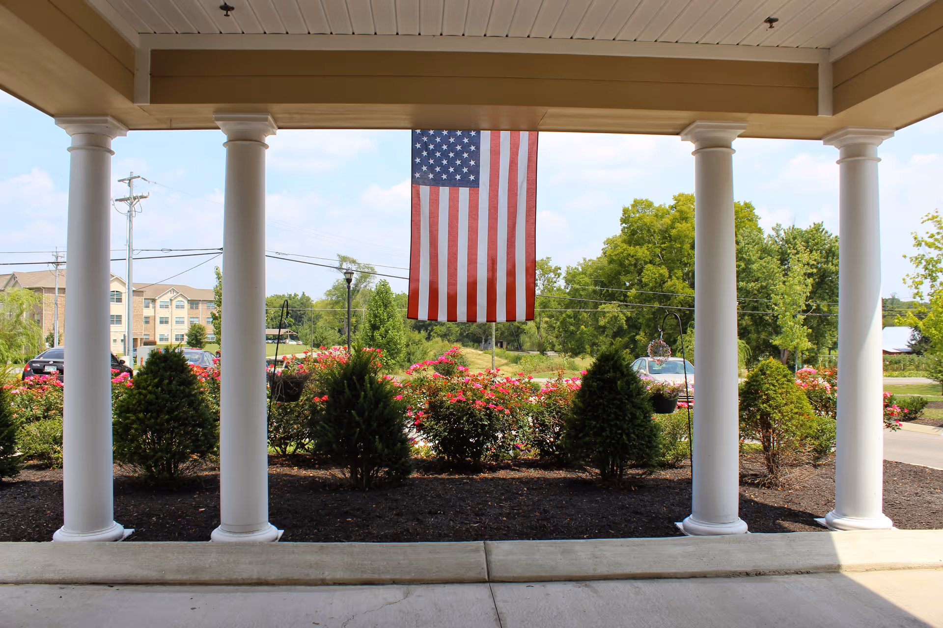 View from a covered porch with white columns looking out onto a landscaped garden with bushes and flowers. An American flag hangs from the porch ceiling. In the background, there are trees, a road, parked cars, and a multi-story building.