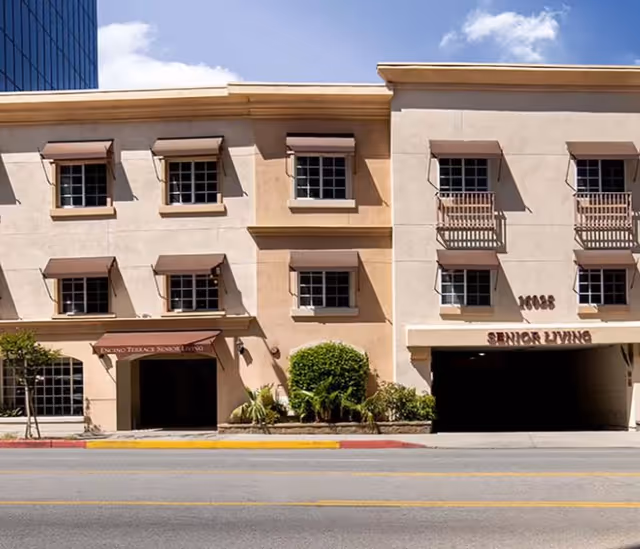 Exterior front view of a three-story senior living facility building with beige walls, multiple windows with awnings, and an entrance labeled Encino Terrace Senior Living. There are small bushes and plants in front of the building along the sidewalk and a clear blue sky above.