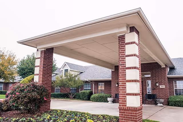 Covered entrance area of a brick building with white trim, surrounded by landscaped bushes and flowers, with a concrete driveway leading up to the entrance.