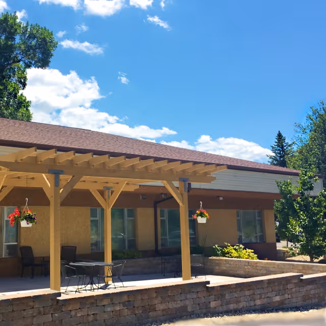 Outdoor patio area at Four Winds Manor with a wooden pergola, hanging flower pots, metal table and chairs, and a low stone wall under a bright blue sky with some clouds.